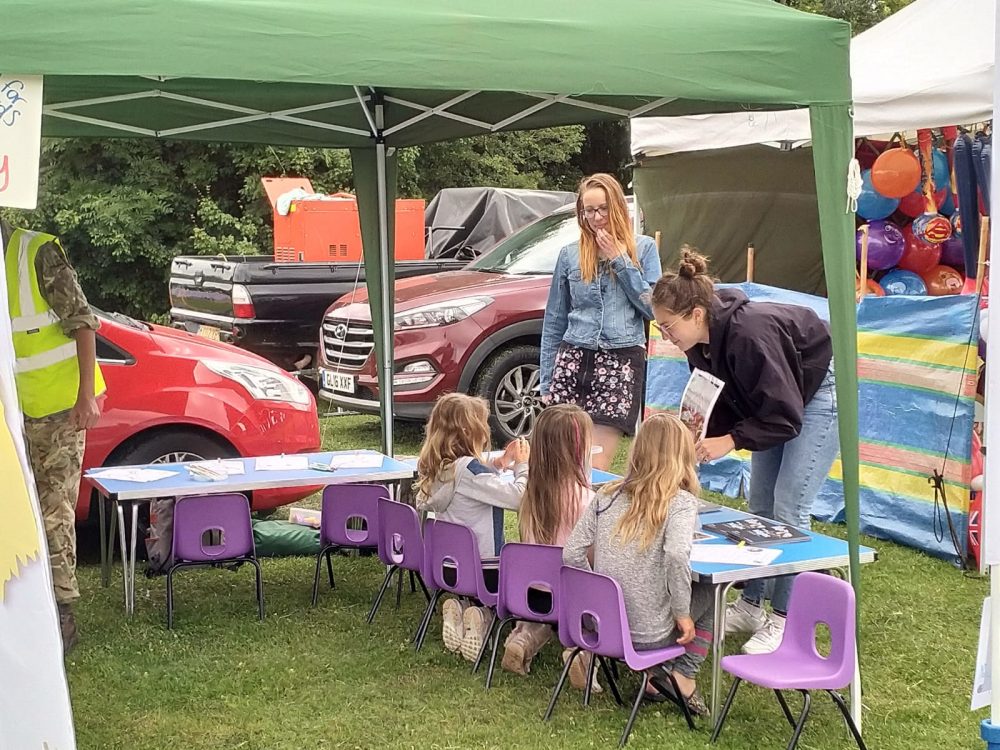 Two young women engaging three children in drawing bats under a marquee at a village fete