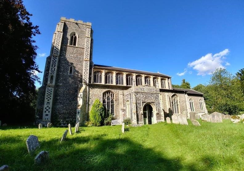 A sunlit church with decorative flint patterning on the buttresses. There are tall, perpendicular windows in the clerestory