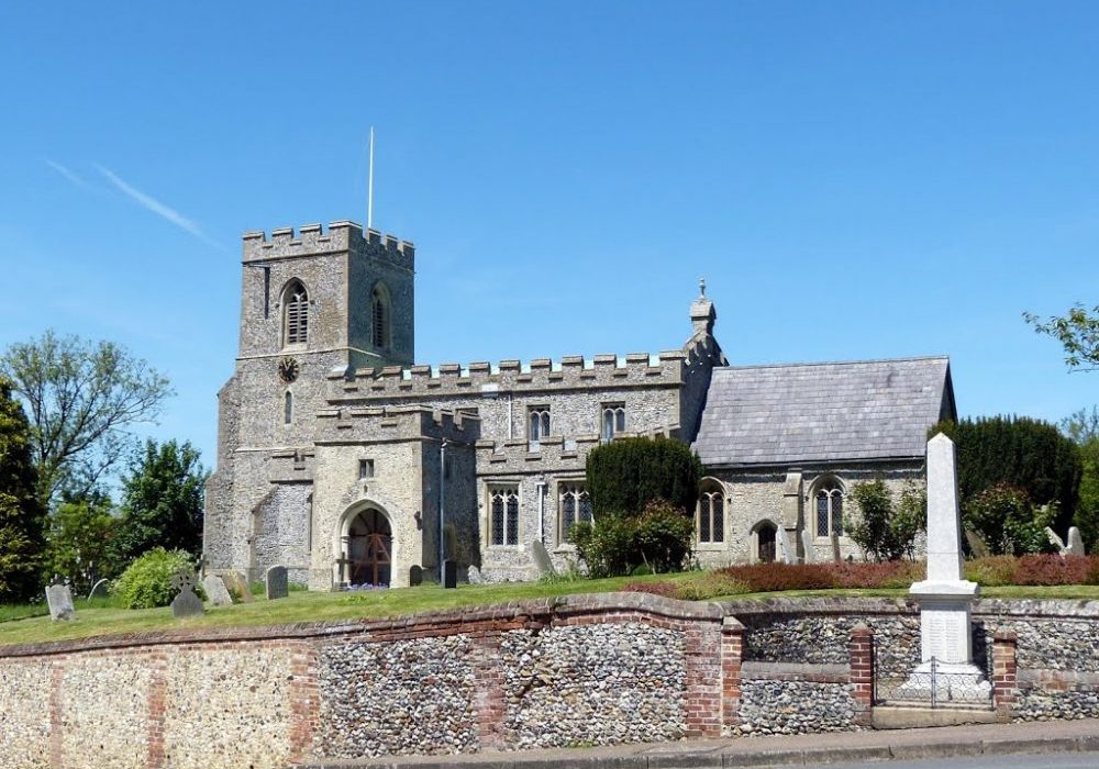 Church exterior, square tower with clock, turreted nave, large porch, flint. Behind flint wall. War memorial