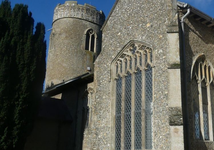 The transept of a flint church from outside, the round tower of the church is just visible in the corner of the image