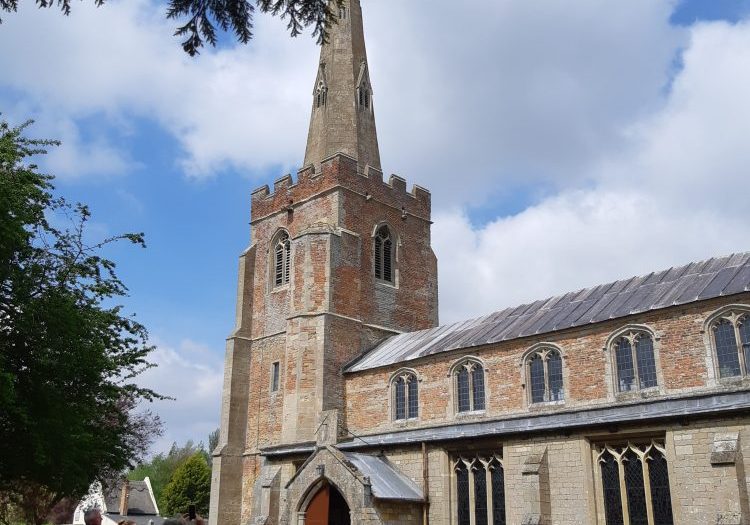 A church made of stone and red brick with a tower topped by a narrow steeple